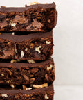 A close-up stack of four thick chocolate brownies with chopped nuts and chocolate chips, shown against a clean off-white background.
