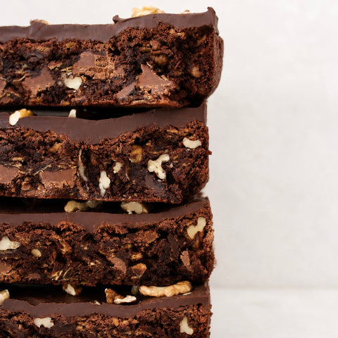 A close-up stack of four thick chocolate brownies with chopped nuts and chocolate chips, shown against a clean off-white background.