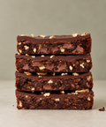 Four stacked chocolate brownies with chopped nuts and chocolate chunks, photographed on a neutral light gray background.