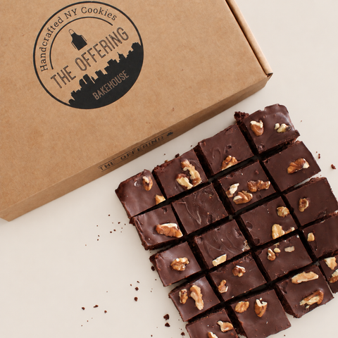 A close-up of chocolate cookie bars topped with chopped walnuts, arranged next to a kraft paper box printed with the brand name 'The Offering Bakehouse'.