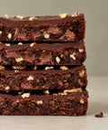 Four stacked chocolate chip cookie bars with a glossy chocolate glaze and chopped nuts, photographed close-up on a neutral light gray background.
