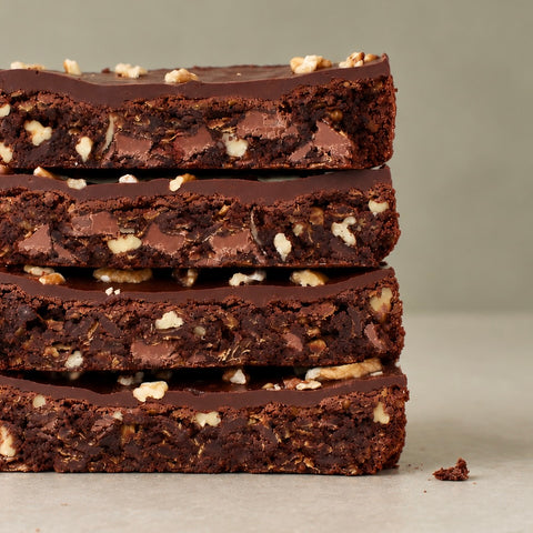 Four stacked chocolate chip cookie bars with a glossy chocolate glaze and chopped nuts, photographed close-up on a neutral light gray background.