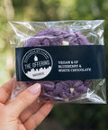 Packaged purple cookies with a clear wrap held by a hand, against a blurred green background.