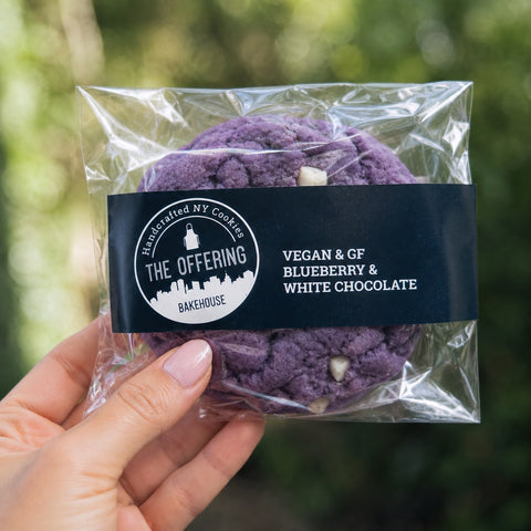 Packaged purple cookies with a clear wrap held by a hand, against a blurred green background.