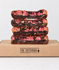 Four stacked chocolate brownies with pink fruit filling and white chocolate, presented on top of a brown cardboard box printed with 'THE OFFERING bakehouse' on a clean white background.