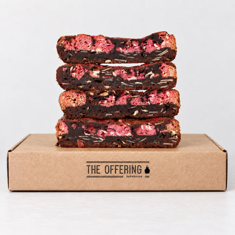 Four stacked chocolate brownies with pink fruit filling and white chocolate, presented on top of a brown cardboard box printed with 'THE OFFERING bakehouse' on a clean white background.