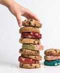 A hand stacks a tall column of assorted cookies, including chocolate chip cookies and colorful red, green, pink, and blue cookies, next to a smaller matching cookie stack on a white background.