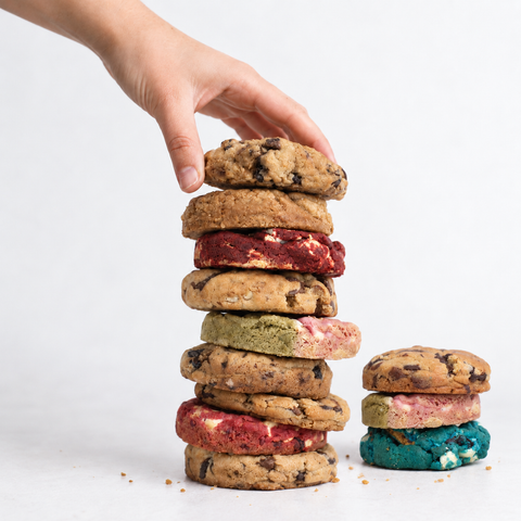 A hand stacks a tall column of assorted cookies, including chocolate chip cookies and colorful red, green, pink, and blue cookies, next to a smaller matching cookie stack on a white background.
