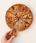 A round chocolate chip cookie pie cut into slices, with one slice being lifted by a hand against a plain light beige background.