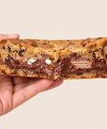 A hand holding a thick chocolate chip cookie or cookie bread slice with melted chocolate and cookie pieces visible in the cross-section, against a plain light beige background.