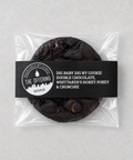 A packaged double chocolate cookie from The Offering Bakery, sealed in a clear plastic bag with a black paper label. The label features white text and a circular logo and is photographed on a neutral light gray background.
