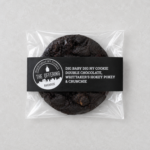 A packaged double chocolate cookie from The Offering Bakery, sealed in a clear plastic bag with a black paper label. The label features white text and a circular logo and is photographed on a neutral light gray background.