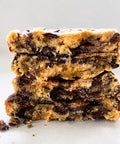 Three stacked chocolate chip cookie bars with melted chocolate chips, photographed close-up on a light neutral background.
