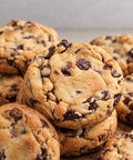 A close-up pile of chocolate chip cookies with golden-brown baked edges and dark chocolate chunks, photographed on a neutral light gray background.