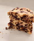 A close-up of a stacked slice of chocolate chip cookie on a light beige background, showing the golden-brown cookie dough and dark chocolate chips in cross-section.