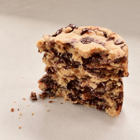 A close-up of a stacked slice of chocolate chip cookie on a light beige background, showing the golden-brown cookie dough and dark chocolate chips in cross-section.