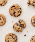 Chocolate chip cookies scattered on a light beige surface, with one cookie broken in half to show the chewy interior.
