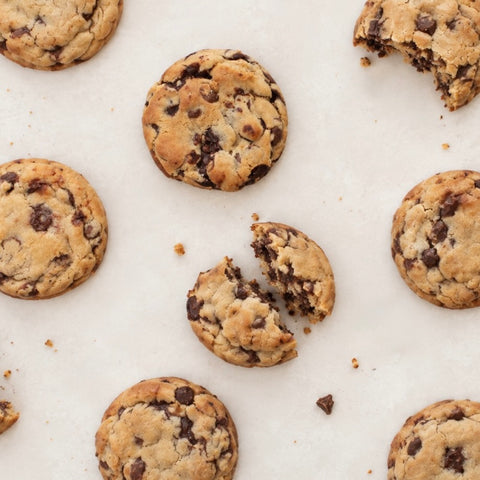 Chocolate chip cookies scattered on a light beige surface, with one cookie broken in half to show the chewy interior.
