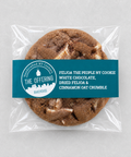 A packaged round cookie on a light gray background. The cookie is sealed in a clear plastic bag with a teal paper label across the front.