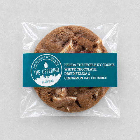 A packaged round cookie on a light gray background. The cookie is sealed in a clear plastic bag with a teal paper label across the front.