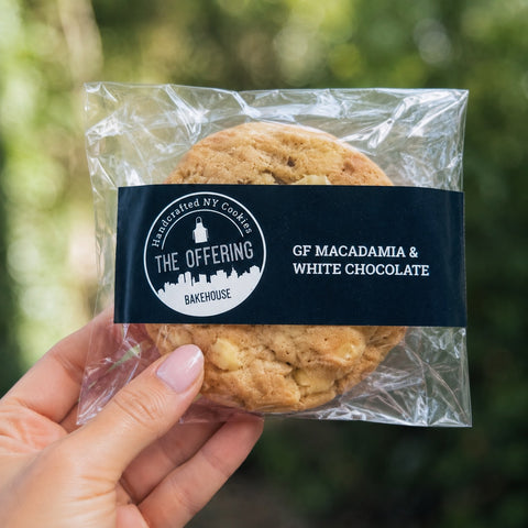 Cookie in clear packaging with 'The Offering Bakehouse' label, held by a hand against a blurred green background.