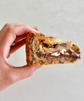 A hand holding a thick slice of chocolate chip cookie with chocolate chunks and a white chocolate drizzle, set against a plain light gray background.