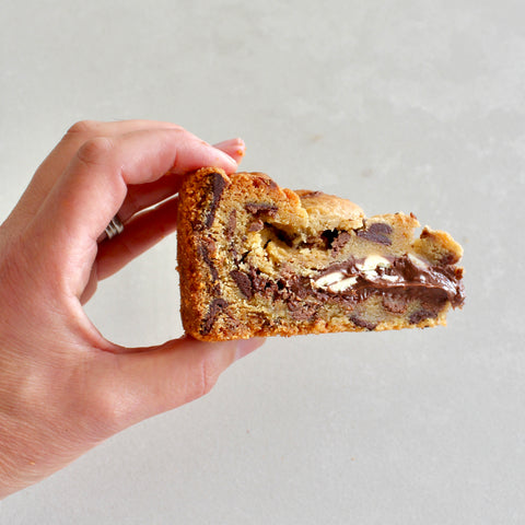 A hand holding a thick slice of chocolate chip cookie with chocolate chunks and a white chocolate drizzle, set against a plain light gray background.