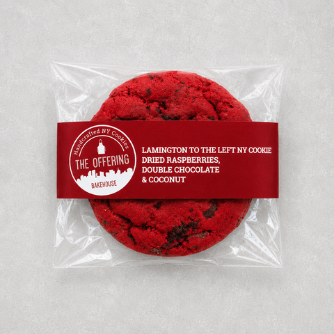 A packaged round cookie on a light gray background. The cookie is sealed in a clear plastic bag with a dark red paper band label.