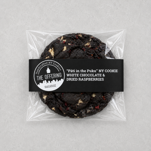 A packaged chocolate chip cookie from The Offering Bakery, featuring a black paper label with white text that reads "Pâté in the Puku" NY COOKIE WHITE CHOCOLATE & DRIED RASPBERRIES. The cookie is visible through a clear plastic bag on a light gray background.