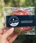 Packaged strawberry matcha and white chocolate cookies held by a hand with a blurred green background