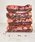 Four stacked chocolate brownies with pink fruit filling and white chocolate drizzle, set on a light gray surface with crumbs scattered around.