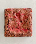 A square brownie with a cracked chocolate surface and bright pink/red berry pieces, photographed from directly above on a light neutral background.