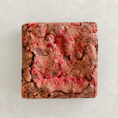 A square brownie with a cracked chocolate surface and bright pink/red berry pieces, photographed from directly above on a light neutral background.
