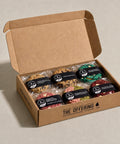 An open kraft cardboard gift box labeled 'THE OFFERING' containing six individually wrapped chocolate chip cookies in assorted flavors, arranged on a light beige surface.