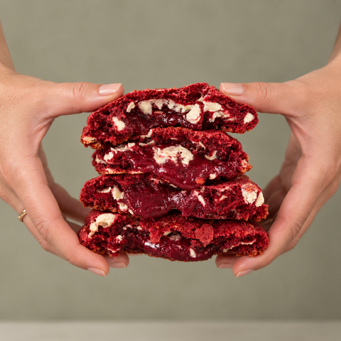 A close-up of two hands holding a stack of four red velvet cookies broken open to show the rich red frosting and white chocolate chunks, set against a solid neutral gray background.