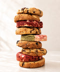 A tall stack of assorted cookies, including chocolate chip cookies, red velvet cookies, and green tea cookie, photographed on a light marble surface with a soft white marble background.