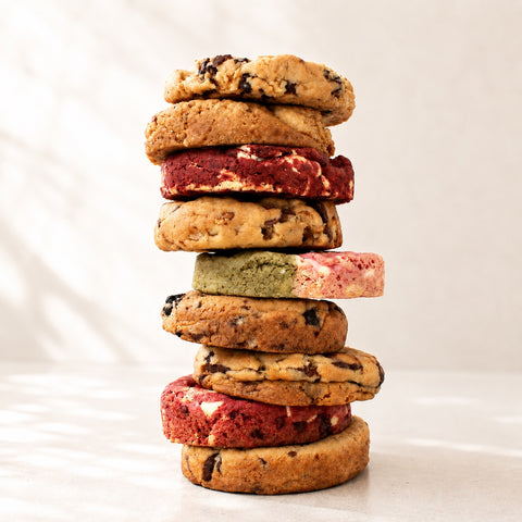 A tall stack of assorted cookies, including chocolate chip cookies, red velvet cookies, and green tea cookie, photographed on a light marble surface with a soft white marble background.