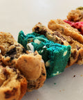 Close-up of assorted cookies arranged diagonally on a light surface, including chocolate chip cookies, a bright blue cookie with white chunks, and a tan cookie with pink filling.