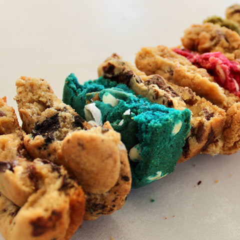 Close-up of assorted cookies arranged diagonally on a light surface, including chocolate chip cookies, a bright blue cookie with white chunks, and a tan cookie with pink filling.