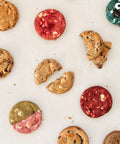 An overhead flat-lay of assorted cookies on a white surface, including chocolate chip cookies, red velvet cookies, and two uniquely decorated cookies with eyes and a pink/red split design.
