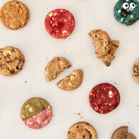 An overhead flat-lay of assorted cookies on a white surface, including chocolate chip cookies, red velvet cookies, and two uniquely decorated cookies with eyes and a pink/red split design.