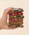 A hand holds a vertical stack of seven colorful baked cookies or blondies against a plain light beige background. The stack features various colored doughs including pink, green, brown, and dark chocolate pieces.