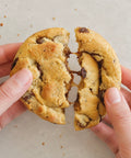 A close-up of a person's hands pulling a chocolate chip cookie apart, showing a stretchy melted chocolate center, on a light neutral background.