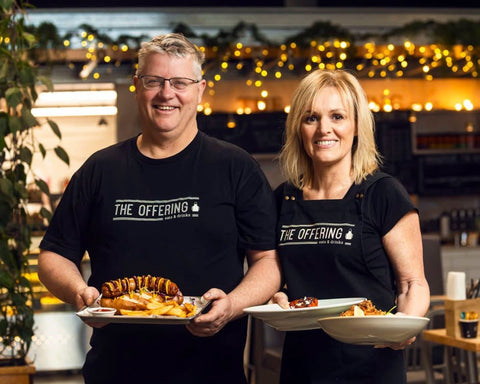 Two people wearing 'The Offering' shirts holding plates of food in a casual setting.