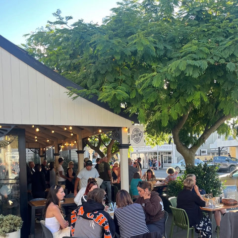 A bustling outdoor patio scene at a restaurant or cafe under a large green tree, with many people seated at tables beside a white-sided structure.
