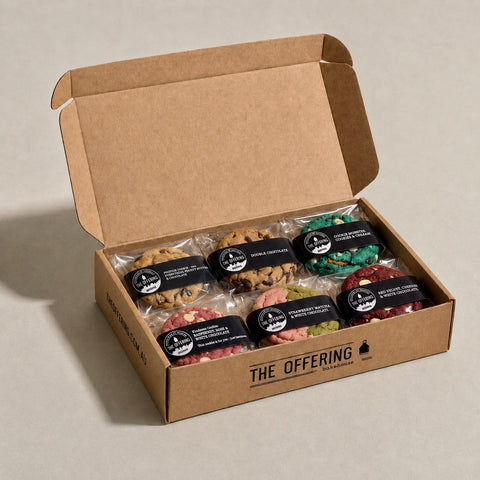 An open kraft cardboard gift box labeled 'THE OFFERING' containing six individually wrapped chocolate chip cookies in assorted flavors, arranged on a light beige surface.
