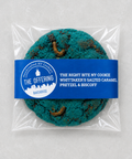 A packaged round cookie in a clear plastic bag with a blue paper label reading 'The Offering' and 'The Right Bite NY Cookie'. The cookie has a bright teal-blue color with brown spots and visible worm-like inclusions, photographed on a light neutral background.