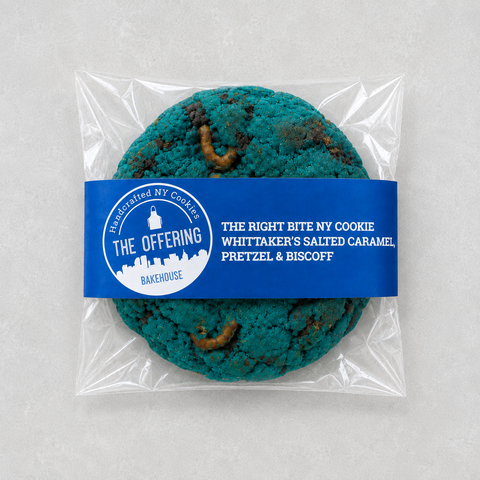 A packaged round cookie in a clear plastic bag with a blue paper label reading 'The Offering' and 'The Right Bite NY Cookie'. The cookie has a bright teal-blue color with brown spots and visible worm-like inclusions, photographed on a light neutral background.