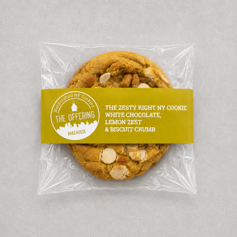 A packaged cookie from The Offering Bakehouse, sealed in clear plastic with a yellow label. The cookie is golden brown with visible white chocolate chips and biscuit crumb pieces.
