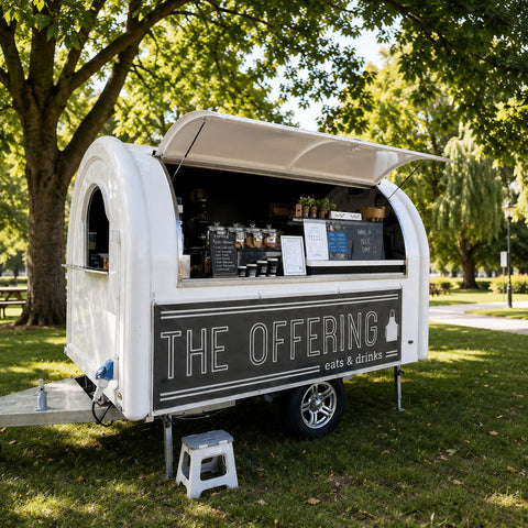 A white mobile food truck with a black side panel reading 'THE OFFERING' and 'eat & drinks' is parked outdoors on grass in a park with large green trees. The serving window is open, showing food items inside, and a small step stool stands in front of the truck.